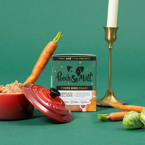 Box of Pooch & Mutt pet food with carrots, a bowl, and a candle on a green background